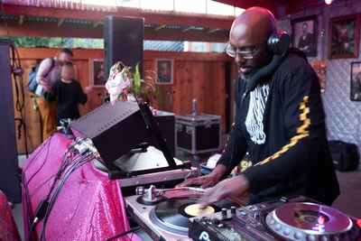DJ Cee White commands the turntables at White Owl Social Club during the season opener of Your Sunday Best day party in Portland, Oregon. Bathed in warm ambient lighting with magenta sequined fabric draped over his DJ equipment, he focuses intently on his vinyl records while wearing headphones and a black graphic tee. The intimate venue's rustic wood-paneled walls adorned with portrait photographs create a cozy backdrop as patrons gather in the softly lit space behind him.