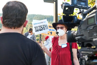 A determined climate activist in a red blouse and black hat addresses KOIN Channel 6 news cameras during a protest at Representative Kurt Schrader's Oregon City office. Behind her, fellow demonstrators hold signs including a phone number urging action, while autumn sunlight filters through the trees of McLoughlin Promenade. The scene captures the intersection of grassroots activism and media coverage, with the activist's white mask and focused demeanor conveying the urgency of climate advocacy in the Pacific Northwest.