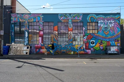 A magnificent large-scale mural transforms an industrial warehouse facade in Portland's Central Eastside district, featuring vibrant psychedelic animals rendered in intricate mandala patterns. The artwork showcases an owl with piercing eyes, a majestic bear, butterflies, and other creatures painted in electric blues, purples, oranges, and greens against the building's teal base. A lone pedestrian walks past the multi-windowed structure, providing human scale to this explosion of street art creativity that epitomizes the neighborhood's artistic renaissance.