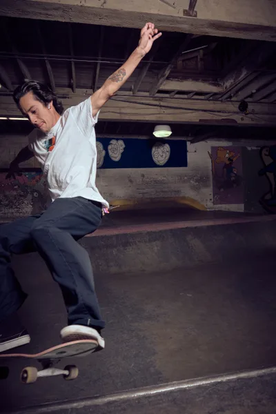 A tattooed skateboarder launches into motion within the gritty confines of Commonwealth Skateboarding's basement venue in Portland's Buckman neighborhood. The raw industrial space, with its exposed wooden beams and graffitied walls, creates a moody backdrop as harsh overhead lighting cuts through the underground atmosphere. The skater's white long-sleeve shirt contrasts sharply against the weathered concrete and dark shadows, while cartoon murals peek through the dimly lit periphery.
