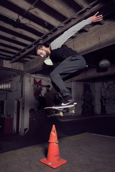 A bearded skateboarder launches into the air above an orange traffic cone in the dimly lit Commonwealth Skateboarding in Portland's Buckman neighborhood. The atmospheric industrial space features exposed wooden ceiling beams and concrete floors, creating a raw underground aesthetic. Moody lighting casts dramatic shadows while graffitied walls and other skaters in the background establish the authentic street culture setting. The frozen moment captures the skater mid-trick, his body extended with arms spread for balance as his board hovers beneath him.