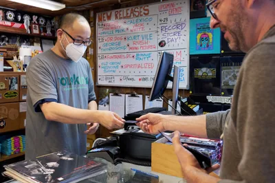 Inside Music Millennium on East Burnside during Record Store Day 2022, a masked clerk in glasses and a gray t-shirt processes a customer's purchase at the checkout counter. The intimate record store setting features handwritten new release boards advertising vinyl from artists like Dr. Dog and Kurt Vile, while shelves packed with albums and merchandise create a cozy backdrop. Warm interior lighting illuminates the exchange between the two men as physical media changes hands in this enduring bastion of music culture during the annual celebration of independent record stores.