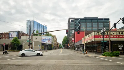 A lone white sedan crosses the eerily deserted intersection of West Burnside Street in Portland's Pearl District, capturing the profound stillness of the early COVID-19 pandemic lockdown. The iconic Powell's Books storefront dominates the right side of the frame, its familiar red and gold signage a stark contrast to the vacant sidewalks that would normally bustle with literary enthusiasts. Overcast skies mirror the somber mood of this historic moment, while modern high-rise towers loom in the background, silent witnesses to the city's sudden pause.