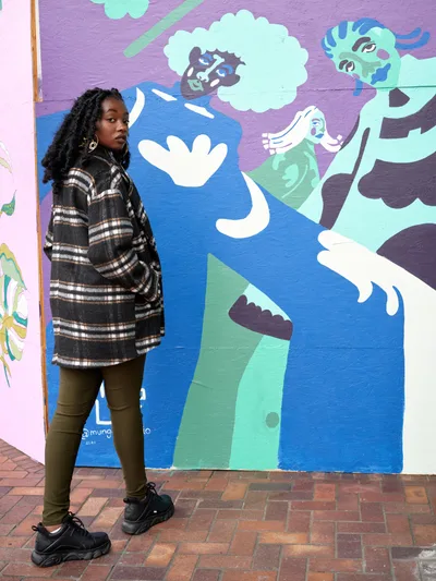 Naomi Likayi stands confidently beside her colorful community mural painted on the boarded Portland World Trade Center, commissioned by the Portland Street Art Alliance. The artist wears a plaid coat and locs, positioned against the whimsical artwork featuring abstract figures in blues, purples, and greens that transform the urban wall into a canvas of hope. Her direct gaze and relaxed stance create an intimate portrait that captures both the artist and her public art contribution to Portland's cultural landscape.