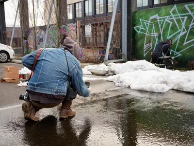 A photographer crouches low to capture a street muralist at work in the parking lot of Clay Creative in Southeast Portland's Hosford-Abernethy neighborhood. The scene unfolds against a backdrop of vibrant graffiti-covered walls, with melting snow creating dark puddles that mirror the overcast winter sky. The intimate composition draws viewers into the creative process, highlighting the dedication required to transform urban spaces into canvases of expression.