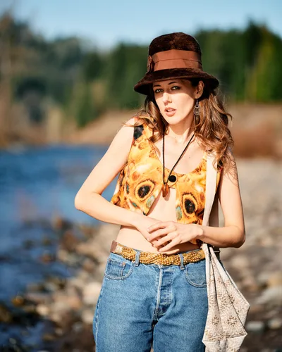 A young woman poses confidently along Oregon's Floodplain Trail, wearing a distinctive butterfly-print vest over a cream top, complemented by high-waisted denim and a russet felt hat with leather band. The warm afternoon light illuminates her flowing brown hair and ornate earrings, while the Columbia River's blue waters and lush Multnomah County landscape create a dreamy, out-of-focus backdrop. Her relaxed stance and gentle expression embody the free-spirited essence of Pacific Northwest outdoor culture.