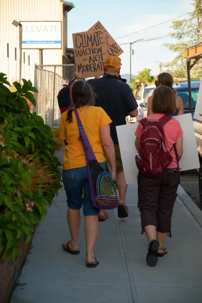 A small group of environmental protesters walks along McLoughlin Promenade in Oregon City, carrying handmade signs advocating for climate action. The late afternoon sun casts long shadows across the concrete sidewalk as the demonstrators make their way past local businesses, including a visible LEVATE sign on a commercial building. The informal procession captures the grassroots nature of climate activism in small-town Oregon, with participants of varying ages united in their call for environmental urgency.