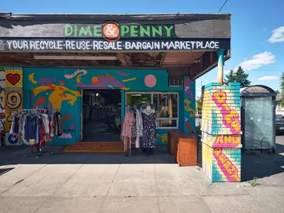 A vibrant thrift shop facade on Northeast 15th and Killingsworth in Portland's Alberta neighborhood bursts with kaleidoscopic murals across weathered walls. Clothing racks spill onto the sidewalk beneath hand-painted signage declaring this a recycle-reuse-resale bargain marketplace. The turquoise, orange, and pink street art transforms the humble storefront into a celebration of sustainable shopping culture.
