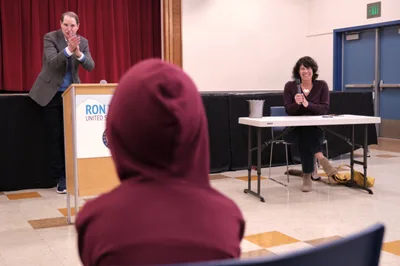 Senator Ron Wyden gestures expressively from behind a podium bearing his name, engaging with constituents during a town hall meeting at Robert Gray Middle School in Portland's Hillsdale neighborhood. The intimate setting features burgundy curtains and polished floors, with a moderator seated at a white table nearby. A community member in a deep red hooded sweatshirt sits in the foreground, representing the engaged local audience participating in this democratic dialogue.