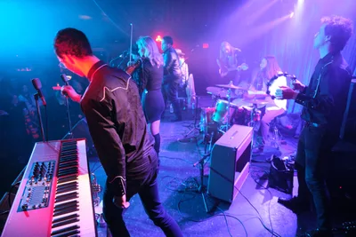 Roselit Bone commands the cramped stage at Portland's legendary Dante's venue, their silhouettes carved by piercing purple and cyan stage lights that slice through the smoky darkness. The band's keyboardist leans into his vintage synthesizer while the guitarist and drummer create a wall of sound behind him, all bathed in the venue's signature neon glow. The intimate basement setting amplifies the electric energy, with cables snaking across the worn floor and amplifiers crowding the small performance space.