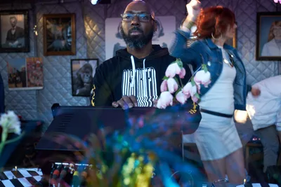 DJ Cee White commands the decks at White Owl Social Club during the season opener of Your Sunday Best day party in Portland, Oregon. Wearing glasses and a black hoodie with white text, he focuses intently on his laptop while a woman in motion blur dances behind him, pink flowers adding color to the intimate venue. The gallery walls adorned with framed artwork create an artistic backdrop for this vibrant Sunday afternoon gathering.