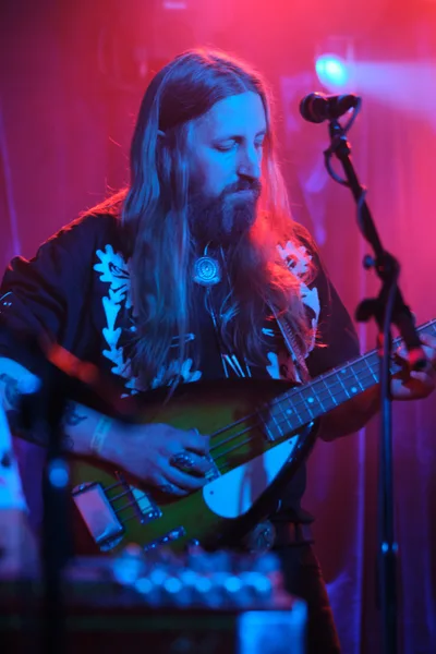 Tobias Berblinger of Roselit Bone commands the stage at Portland's legendary Dante's venue, his flowing blonde hair catching the saturated magenta and cyan stage lights. The bassist stands in contemplative focus, adorned in a black band tee and pendant necklace, while the atmospheric concert lighting transforms the intimate club into an otherworldly sonic cathedral. The image captures a moment of musical transcendence during RX Fest 2019, where underground music culture thrives in Portland's creative heart.