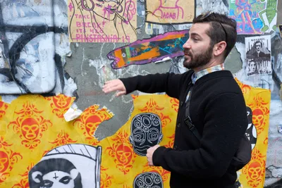 Tomás Valladares gestures enthusiastically toward a weathered concrete wall layered with vibrant street art, wheat paste installations, and graffiti in Portland's Central Eastside district. The bearded guide, dressed in a dark sweater, stands before a kaleidoscope of urban expression featuring yellow skull motifs, colorful paste-ups, and layered artwork that tells the story of the neighborhood's creative pulse. Behind him, the textured wall serves as a living gallery where artists have built upon each other's work, creating a palimpsest of contemporary street culture.