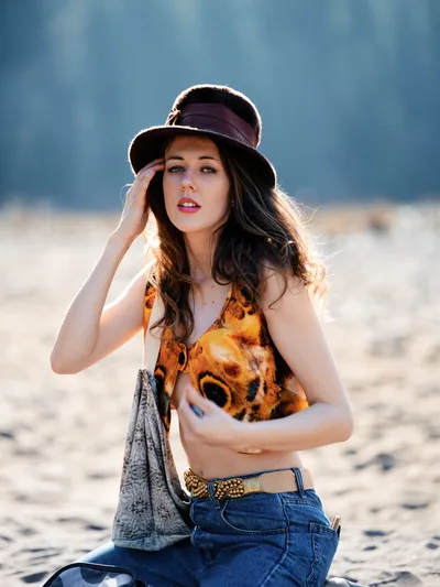 A young woman with flowing dark hair poses thoughtfully in the warm golden light of late afternoon along Oregon's Floodplain Trail in Multnomah County. She wears a burgundy wide-brimmed hat and vibrant leopard print top paired with high-waisted denim, her hand gracefully touching her temple in a contemplative gesture. The soft backlighting creates a luminous rim around her silhouette against the hazy blue-gray backdrop of the Columbia River Gorge landscape.