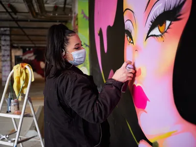 Multidisciplinary artist Vilatose meticulously applies spray paint to a luminous female portrait mural within the industrial confines of Clay Creative's parking garage in Portland's Hosford-Abernethy neighborhood. Wearing a protective mask and dark hoodie, the artist works with focused precision on the dramatic face featuring amber eyes and flowing black hair rendered in electric pinks, purples, and coral hues. The raw concrete space transforms into an urban canvas, with paint supplies and a stepladder nearby suggesting hours of dedicated creative labor.