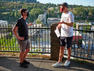 Michael Gaskill and Peter Marks engage in earnest conversation during a climate change protest at Representative Kurt Schrader's office in Oregon City. Standing against black iron railings with the Willamette River valley and forested hills stretching behind them, both men wear face masks in the golden afternoon light. The intimate moment captures the grassroots nature of political activism, with Marks holding papers while gesturing toward his companion against the backdrop of the McLoughlin Promenade overlook.