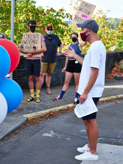 Organizer Peter Marks gestures emphatically while speaking to a small crowd of masked protesters gathered outside Representative Kurt Schrader's office in Oregon City. The speaker, wearing a white t-shirt and baseball cap, holds papers while addressing activists holding signs reading "Climate Inaction Not Modest" and "Corporate Welfare is Not Science." Autumn foliage creates a warm backdrop against the urban setting of McLoughlin Promenade, capturing a moment of grassroots political engagement during the pandemic era.