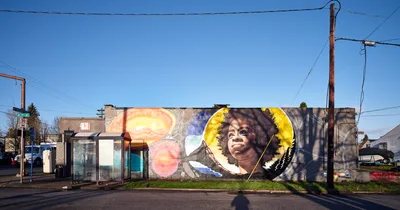 A vibrant street mural depicting poet Amanda Gorman adorns the side wall of a commercial building at the intersection of Northeast 15th and Killingsworth in Portland's Alberta district. The artwork features Gorman's portrait surrounded by golden celestial imagery and cosmic elements, rendered in warm yellows and cool blues against the clear winter sky. Power lines crisscross overhead while bare tree branches frame the scene, creating an urban intersection where art transforms everyday infrastructure into a celebration of literary achievement.