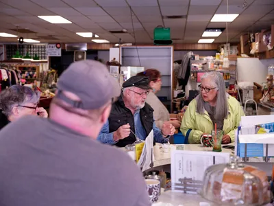 Under the warm glow of fluorescent ceiling lights, patrons gather at the worn formica counter of Pattie's Home Plate Café in Portland's Saint Johns neighborhood. A bearded man in a black cap and vest leans forward in animated conversation with a woman in a bright yellow jacket, her long gray hair catching the light as she gestures expressively. The intimate counter scene captures the timeless ritual of neighborhood dining, where scattered newspapers, coffee cups, and the cluttered backdrop of a working kitchen create an authentic portrait of blue-collar community life.