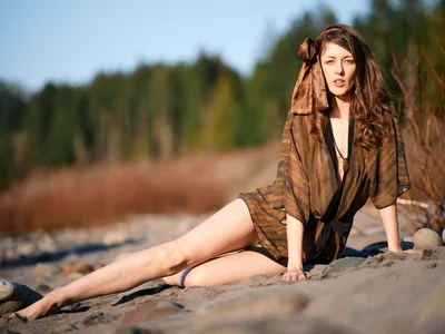 A young woman reclines gracefully on weathered driftwood along the sandy shores of an Oregon riverbed in Multnomah County. Draped in an earthy, patterned kimono that catches the amber light of golden hour, she gazes contemplatively toward the camera while dense Pacific Northwest forest creates a soft, verdant backdrop. The warm, directional lighting sculpts her features and illuminates the natural textures of sand, wood, and flowing fabric.