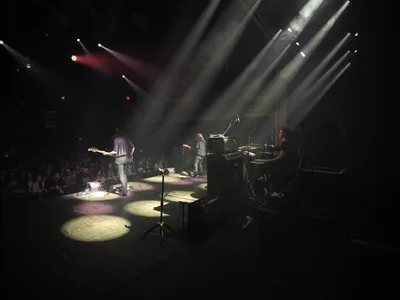 Emma Richardson, Julian Dorio, and Russell Marsden of Band of Skulls perform during their 10 Year Anniversary Tour at Manhattan's historic Webster Hall. Dramatic stage lighting cuts through the venue's darkness, casting golden pools across the performance space while silhouetting the trio against towering light beams. The intimate concert setting captures the raw energy of live rock performance, with the audience gathered in shadow around the illuminated stage.