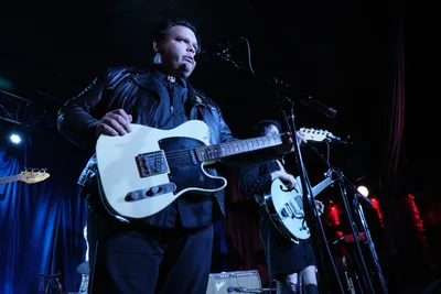 Victor Franco of Roselit Bone grips his white Telecaster guitar under dramatic blue stage lighting at Dante's in Portland, Oregon during RX Fest 2019. The leather-jacketed performer stands focused at the microphone, his intense gaze cutting through the moody concert atmosphere while bandmates remain partially visible in the shadowed background. The intimate venue's low ceiling and close quarters create an electric tension between artist and audience.