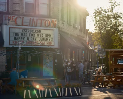 The exterior of The Clinton Street Theater in Portland, Oregon, bathed in warm golden lens-flare sunlight. The theater's marquee reads "The Mad Fox Wed 8PM — Happy B Day Jeremy!! — Rocky Horror Sat Mid." A lively crowd of people gathers on the sidewalk outside, with outdoor picnic tables and planters with marigolds occupying a parklet in the foreground. A woman in a face mask wearing a black "Agnès Varda Forever" t-shirt walks past. Bicycles are parked along the curb, and a woman sits alone at a picnic table to the right.