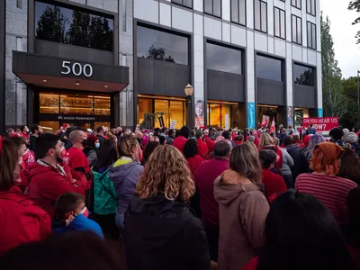 A sea of red-clad healthcare workers fills the sidewalk outside Kaiser Permanente's modernist headquarters at 500 Northeast Multnomah Street in Portland's Lloyd District. The crowd of striking nurses creates a powerful visual contrast against the building's sleek glass facade and concrete columns, their unified presence illuminated by the warm glow spilling from the medical center's ground-floor windows in the gathering dusk. Signs demanding "CAN YOU HEAR US NOW?" punctuate the peaceful but determined assembly, while the institutional architecture looms overhead, emphasizing the David-versus-Goliath nature of the labor dispute.