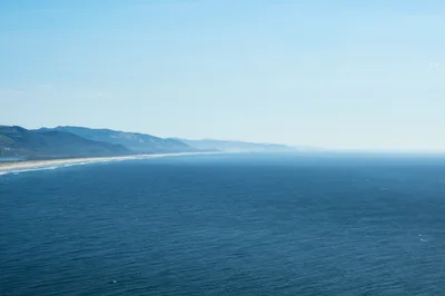 An expansive aerial view captures the Oregon coast where the Pacific Ocean meets the rugged Tillamook County shoreline. The deep blue waters stretch endlessly toward a hazy horizon, while forested mountains roll inland from a pristine sandy beach. Soft atmospheric haze creates layers of blue tones that blend sea, sky, and distant peaks into a seamless coastal panorama.
