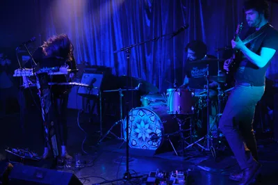Sun Blood Stories delivers an atmospheric performance bathed in ethereal blue and purple stage lighting at Dante's in downtown Portland. The trio creates an intimate soundscape with synthesizers, drums, and guitar, their silhouettes emerging from the moody wash of color against draped curtains. The geometric mandala design on the drum kit serves as a focal point amid the sea of electronic equipment and cables sprawling across the venue floor.