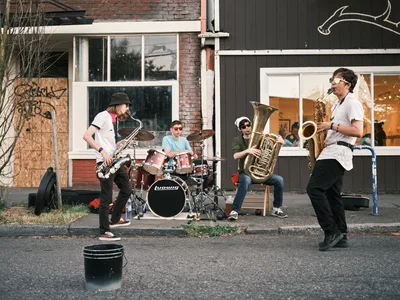 Four young musicians perform an impromptu jazz session on the weathered asphalt of Northeast Alberta Street during Portland's Last Thursday art walk. Golden afternoon light bathes the scene as a saxophonist in a black beanie leads the ensemble, while a drummer, tuba player, and second saxophonist create an intimate street corner concert. Behind them, the characteristic mix of brick and dark wood siding reflects the gentrifying neighborhood's evolving character, with curious onlookers visible through storefront windows.