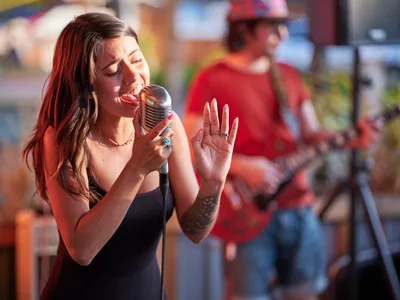 Aubrey Gordon leans into her vintage microphone with passionate intensity, her eyes closed and hand raised expressively as golden late-afternoon light bathes her performance at Portland Mercado. The warm sunlight catches her auburn hair and black sleeveless top while a blurred musician in red provides depth behind her. The intimate outdoor venue creates a compelling contrast between the sharp focus on Gordon's emotional delivery and the softly rendered background of Southeast Portland's vibrant food cart pod.