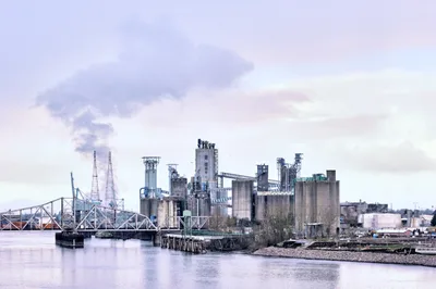 The United Grain Corporation's Vancouver Export Terminal rises from the Columbia River's edge in a symphony of concrete silos, steel conveyor bridges, and industrial towers. Captured during the warm afternoon hours, the facility's imposing structures are bathed in soft lavender and rose-tinted light, their reflections shimmering across the glassy water. A steel truss bridge spans the river in the left frame, connecting this vital grain export hub to the broader Pacific Northwest transportation network.