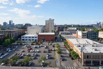An elevated view captures Portland's historic Chinatown district on a bright afternoon, where brick facades and contemporary structures create an eclectic urban tapestry. The bustling street scene unfolds below with diagonal parking lots filled with vehicles, tree-lined sidewalks, and pedestrians navigating the intersection of heritage and modern commerce. Puffy white clouds drift across azure skies above this Northwest Portland neighborhood, while the distinctive architecture tells the story of cultural evolution in the Pacific Northwest.