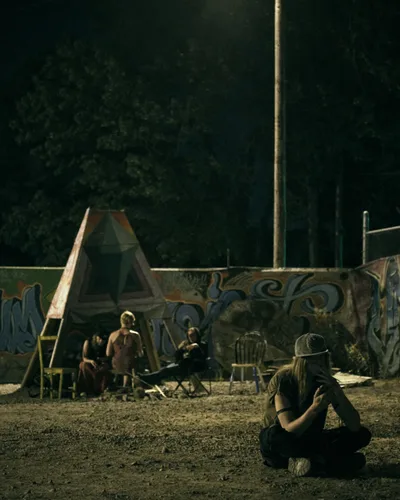 Under the amber glow of a single street lamp, members of a Portland art collective gather in their outdoor space on Southeast 76th Avenue. A man with long hair and baseball cap sits cross-legged in the foreground, while shadowy figures socialize among art installation structures and graffiti-adorned walls. The nocturnal scene captures the community spirit of Mount Tabor's underground creative collective, where artists connect in the darkness between day and dawn.