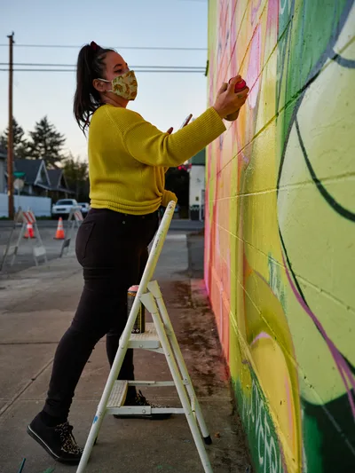 Multidisciplinary artist Villatose balances on a white stepladder, applying vivid streaks of pink and yellow paint to her towering wall mural 'Jardín Cubana' in Portland's Alberta Arts District. The artwork depicts three female figures rendered in flowing brushstrokes across the concrete canvas. Dressed in a mustard-yellow ribbed sweater and floral face mask, she works with focused determination as twilight descends over Northeast Alberta Street. The rainbow cascade of colors flows down behind her, while power lines and modest neighborhood buildings frame this moment of creative transformation in one of the city's most celebrated artistic corridors.