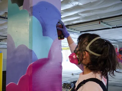 A young muralist wearing a protective respirator mask guides a spray paint canister across a concrete pillar, layering vibrant blues and magentas in flowing abstract forms. The industrial setting of Clay Creative's parking garage in Southeast Portland frames her focused work, with corrugated metal ceiling panels casting diffused light over the evolving artwork. Her concentrated posture and deliberate movements capture the meditative intensity of urban art creation in progress.
