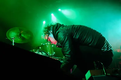 Electronic musician Eli Savage, performing as Ellipsism, hunches intensely over his equipment during a live set at RXFest 2019 in Portland's legendary Dante's venue. Bathed in ethereal green stage lighting that cuts through billowing fog, the artist's concentrated posture and wild curls create a dramatic silhouette against the luminous backdrop. The atmospheric haze and piercing spotlights transform the intimate club space into an otherworldly sonic landscape, with drum kit and electronic gear barely visible in the moody chiaroscuro.
