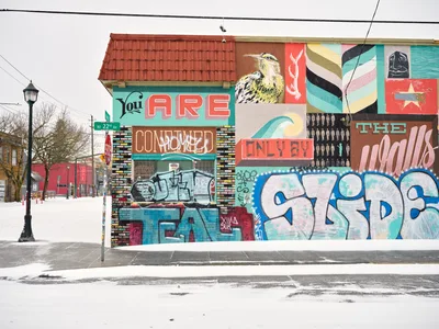 A vibrant community mural by Blaine Fontana dominates a brick building on Northeast Alberta Street in Portland's Alberta Arts District, its colorful proclamation "You ARE" anchoring a collage of street art elements including a majestic bird, geometric patterns, and layered graffiti tags. Fresh snow blankets the quiet sidewalk and street, creating a stark contrast against the warm reds of the clay tile roof and the explosion of turquoise, coral, and pink hues across the wall. The overcast winter sky and vintage street lamp complete this portrait of Portland's celebrated public art scene, where commissioned murals coexist with spontaneous street expression.