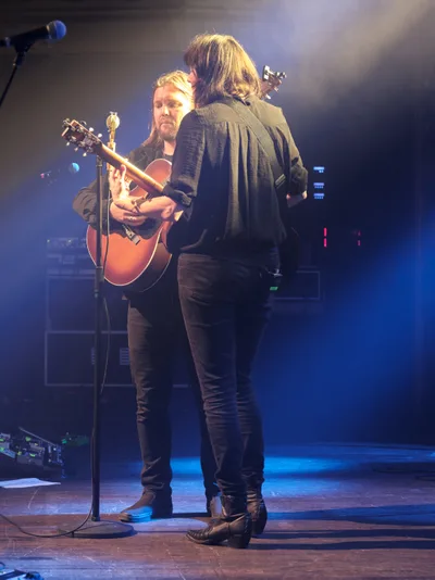 Emma Richardson and Russell Marsden of Band of Skulls share an intimate musical moment during their 10 Year Anniversary Tour at Webster Hall in Manhattan. Bathed in ethereal blue stage lighting with atmospheric haze, Richardson stands with her back to the audience while Marsden faces forward, cradling an acoustic guitar at the microphone. The dramatic lighting creates a moody, contemplative atmosphere that captures the emotional weight of their decade-long musical journey.