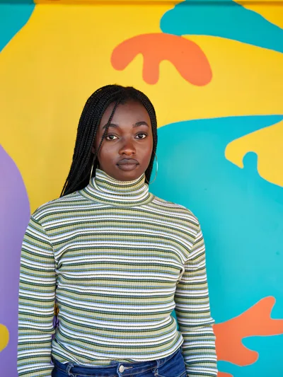 Naomi Likayi stands confidently before her vibrant street mural in Portland's Alberta district, wearing a striped turtleneck that echoes the rhythmic patterns of her artistic work. The kaleidoscopic backdrop bursts with flowing shapes in turquoise, golden yellow, coral, and purple, creating an energetic conversation between the artist and her creation. Soft, even lighting illuminates her direct gaze and the intricate box braids that frame her face, while the organic forms of the mural seem to dance around her figure.