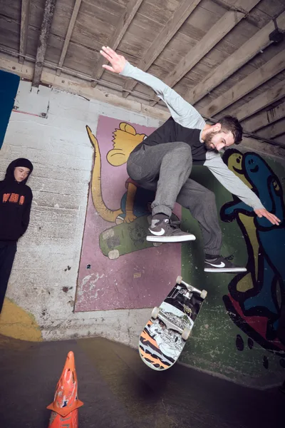 A bearded skateboarder executes a dynamic aerial maneuver in the concrete bowl at Commonwealth Skateboarding in Portland's Buckman neighborhood. The raw industrial space features exposed wooden beams overhead and colorful murals painted directly on the bowl's surfaces, including whimsical cartoon characters that contrast with the gritty concrete architecture. Another skater in a black hoodie observes from the sidelines as late afternoon light filters through the warehouse, creating dramatic shadows across the worn skating surfaces.