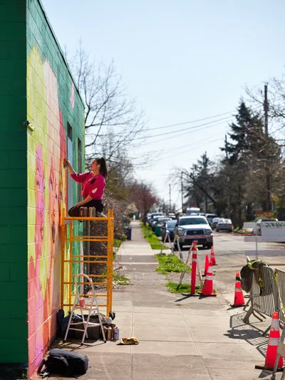 Multidisciplinary artist Vilatose perches on an orange scaffolding platform, painting vibrant geometric patterns onto the side wall of a building along Northeast Alberta Street in Portland's Alberta Arts District. The artist, wearing a bright pink sweatshirt, works methodically on the colorful mural featuring bold blocks of yellow, green, and coral against the overcast spring sky. Orange traffic cones line the sidewalk below, marking the creative workspace as neighborhood traffic flows past on the tree-lined street.