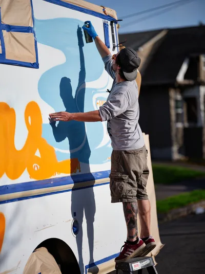A tattooed muralist in a backwards cap and grey sweatshirt stretches upward with spray paint, adding vibrant orange and blue lettering to the side of a white food truck. Golden afternoon light illuminates his focused work as his shadow falls dramatically across the vehicle's surface. The residential Portland neighborhood setting creates an intimate backdrop for this moment of urban artistry in progress.