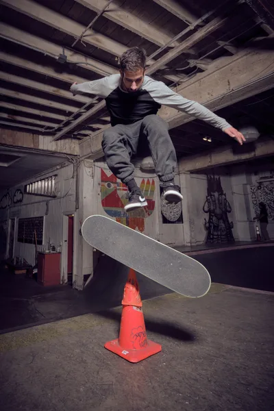 A young skateboarder executes a precise trick over an orange traffic cone in the raw, industrial confines of Commonwealth Skateboarding in Portland's Buckman neighborhood. Exposed wooden ceiling joists and graffiti-adorned concrete walls create a gritty backdrop, while dramatic lighting captures the athlete suspended mid-air above the improvised obstacle. The weathered concrete floor and urban decay aesthetic embody the authentic underground skateboarding culture that thrives in repurposed spaces.