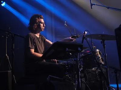 Drummer Julian Dorio of Band of Skulls sits poised behind his kit at Webster Hall during the band's 10 Year Anniversary Tour, bathed in ethereal blue stage lighting that cuts through the intimate venue's signature darkness. His focused expression and relaxed posture capture a moment of quiet intensity between songs, while overhead lighting rigs and microphone stands frame the scene. The atmospheric haze and dramatic chiaroscuro lighting transform the historic Manhattan music hall into a cathedral of sound.