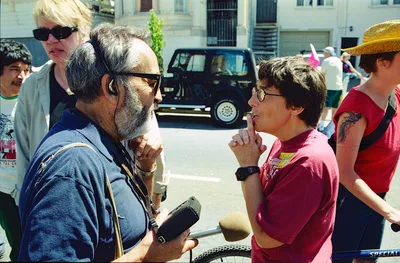 In the sun-drenched plaza at Bryant Street, a bearded journalist conducts an intimate street interview with a local activist during a Mission Anti-Displacement Coalition demonstration. The reporter leans forward intently, microphone extended, while his subject speaks passionately about gentrification's impact on San Francisco's historic Latino neighborhood. Behind them, fellow protesters gather in the harsh midday light, their presence adding urgency to this moment of grassroots media coverage.