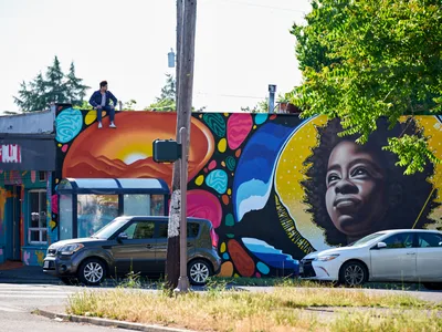 Muralist Travis Fields sits triumphantly atop the completed rooftop of a building adorned with his vibrant portrait of poet Amanda Gorman in Portland's Alberta district. The expansive mural features Gorman's face surrounded by a golden halo and kaleidoscopic patterns of turquoise, magenta, and orange that cascade across the building's facade. Two parked cars rest beneath the artwork while lush green foliage frames the scene under an overcast Pacific Northwest sky.