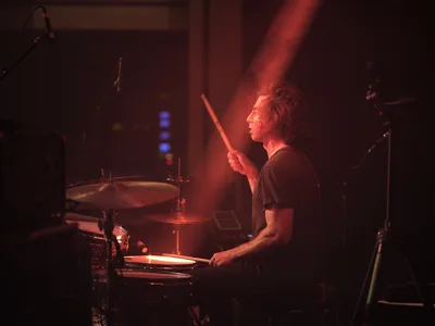 Julian Dorio, drummer for Band of Skulls, sits bathed in dramatic red stage lighting during the band's 10 Year Anniversary Tour at Webster Hall in Manhattan. The atmospheric concert photography captures him mid-performance, drumsticks raised, his profile illuminated against the moody crimson glow that defines the intimate venue's legendary ambiance. Equipment and microphone stands frame the composition in the shadowy background.