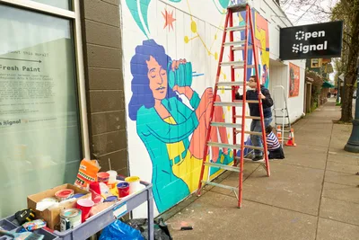 Artists Anke Gladnick and Maria Rodriguez collaborate on a vibrant community mural titled 'Let's Talk' outside Open Signal in Portland's Eliot neighborhood. The scene captures the creative process in action, with a red ladder positioned against the building wall where a colorful figure with flowing blue hair emerges from fresh paint. Paint cans and supplies scattered on a makeshift table reveal the organized chaos of artistic creation, while the 'Open Signal' sign overhead anchors the cultural significance of this Northeast Martin Luther King Jr. Boulevard location.